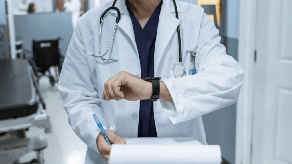 A doctor in a white lab coat and stethoscope checking his watch while holding a clipboard in a hospital hallway.