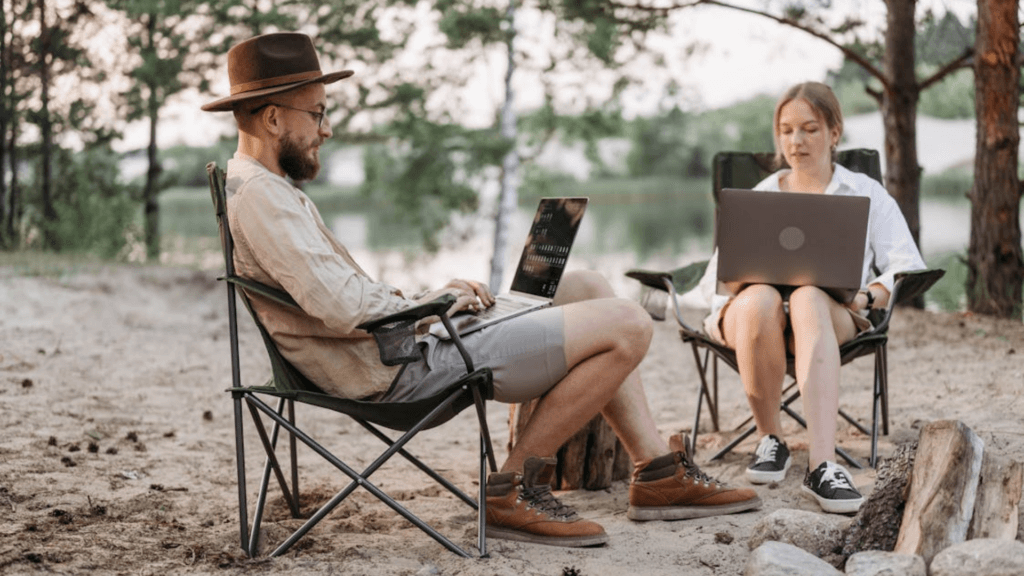 A man and a woman sit in folding chairs on a sandy beach both working on laptops near a small campfire.