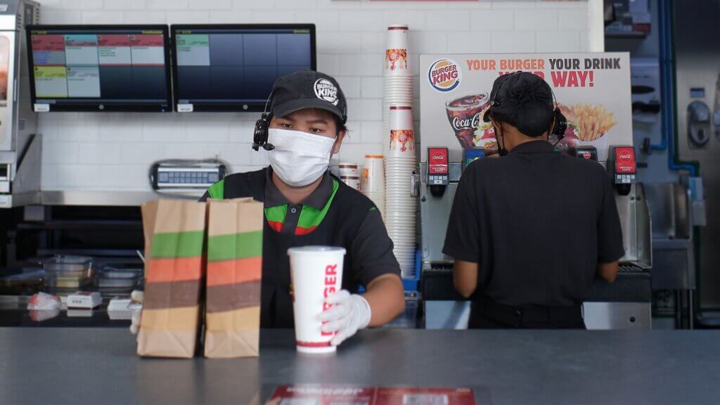 A Burger King employee wearing a headset and mask holding a drink and two paper bags at the counter while another worker is at the back.