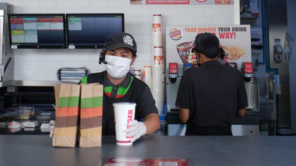 A fast-food worker wearing a mask and gloves serves a drink next to two paper bags. Another worker in the background fills a cup at a soda machine.