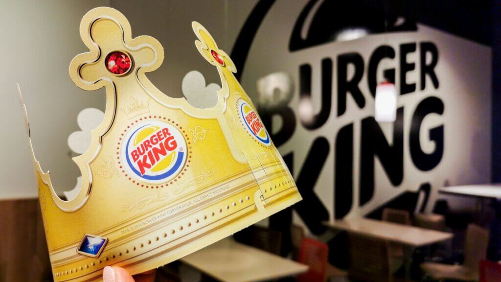 A hand holds a paper Burger King crown decorated with colored gems. In the background, a blurred Burger King logo is visible inside the restaurant.