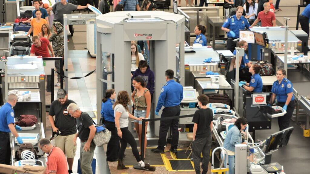 Passengers placing bags on conveyor belts at airport security screening with TSA officers monitoring lines.