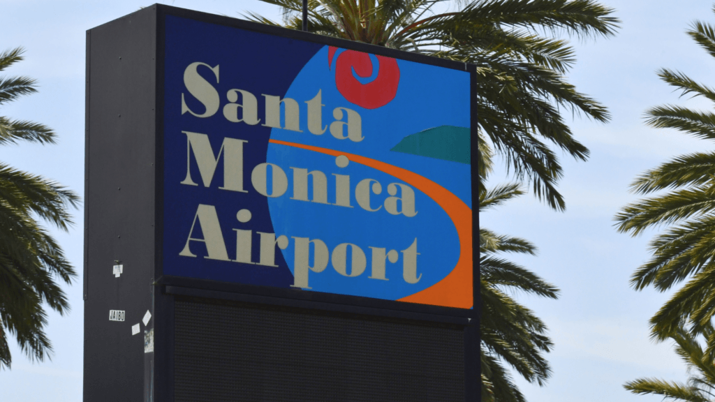 A close-up of a blue and orange roadside monument sign that reads _Santa Monica Airport_ surrounded by palm trees.
