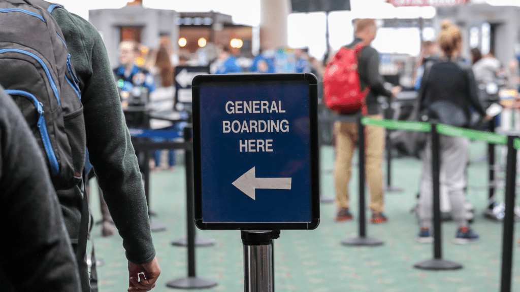 A close-up of a blue directional sign at an airport that reads _GENERAL BOARDING HERE_ with a white arrow pointing to the left.