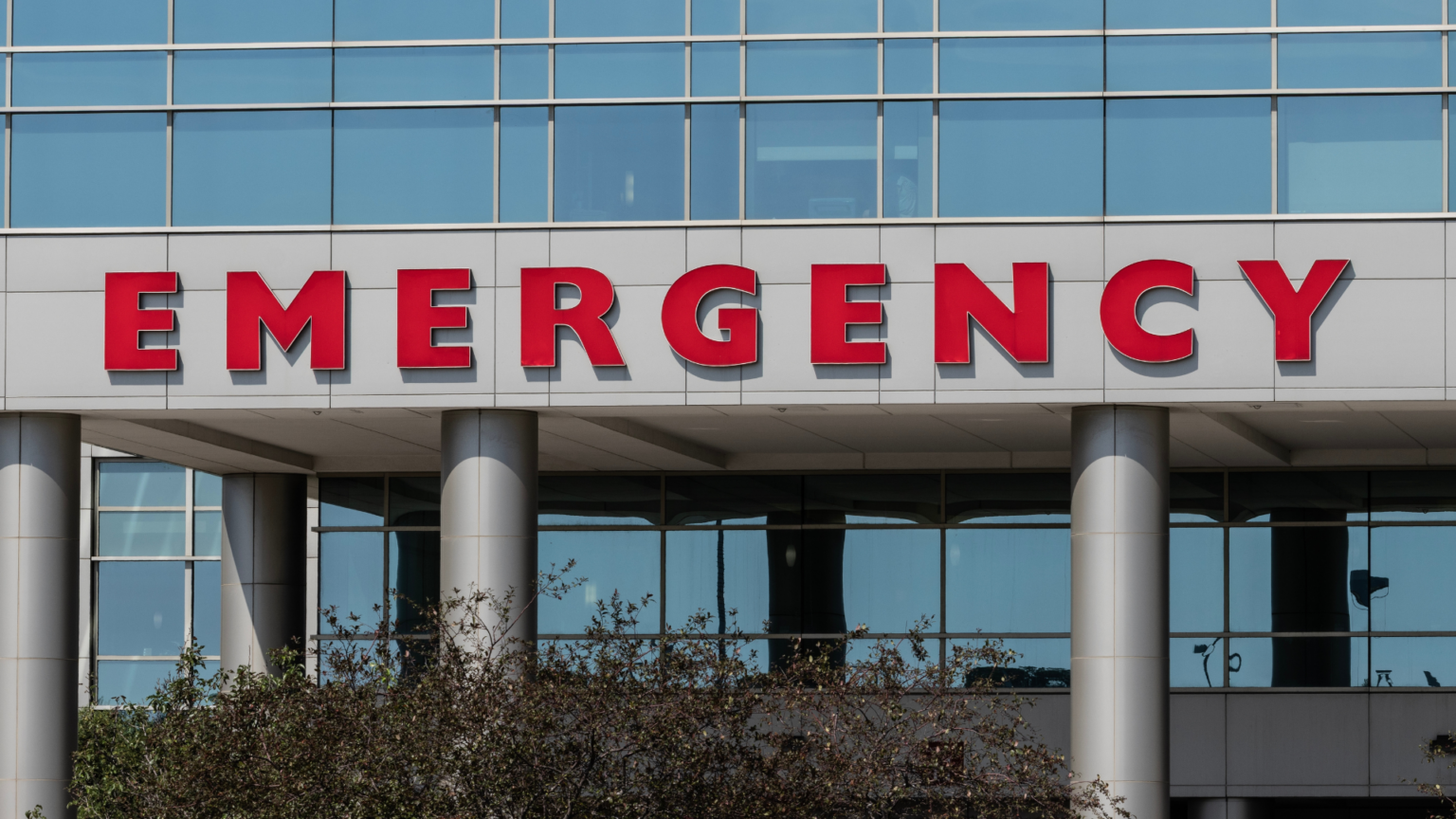 A large, red _EMERGENCY_ sign on the exterior of a modern glass hospital building.