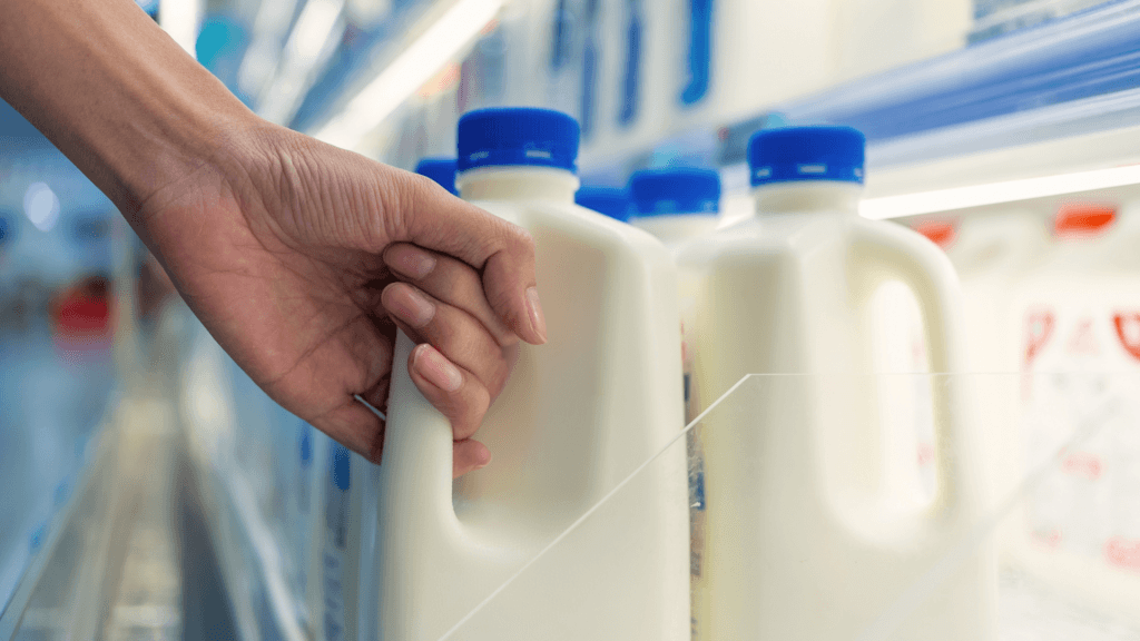 A person's hand reaching for a plastic gallon jug of milk on a refrigerated grocery store shelf.