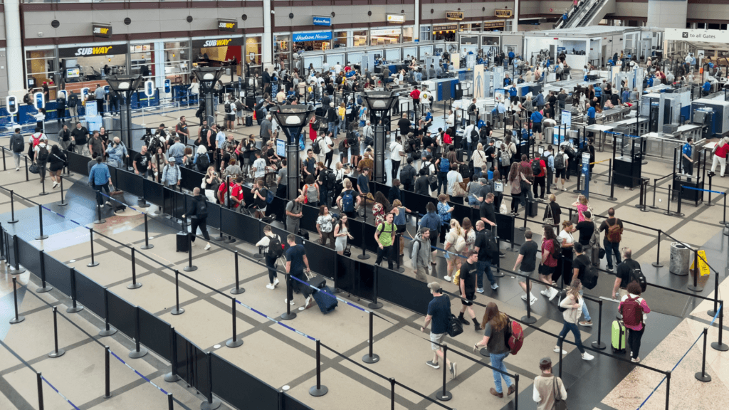 A wide aerial view of a bustling airport security checkpoint with long lines of travelers winding through tensa-barriers in a large terminal with high, white arched ceilings.