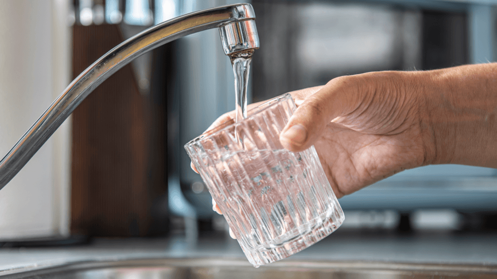 Closeup of a glass help by a hand being filled with tap water in a sink.