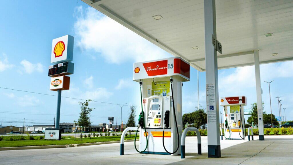 A bright daytime view of a Shell gas station with multiple fuel pumps under a canopy. The Shell logo is visible on signs and pumps.