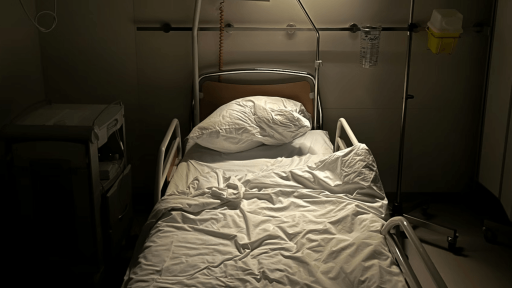 A dimly lit hospital room showing an empty bed with rumpled white sheets and medical equipment standing nearby.