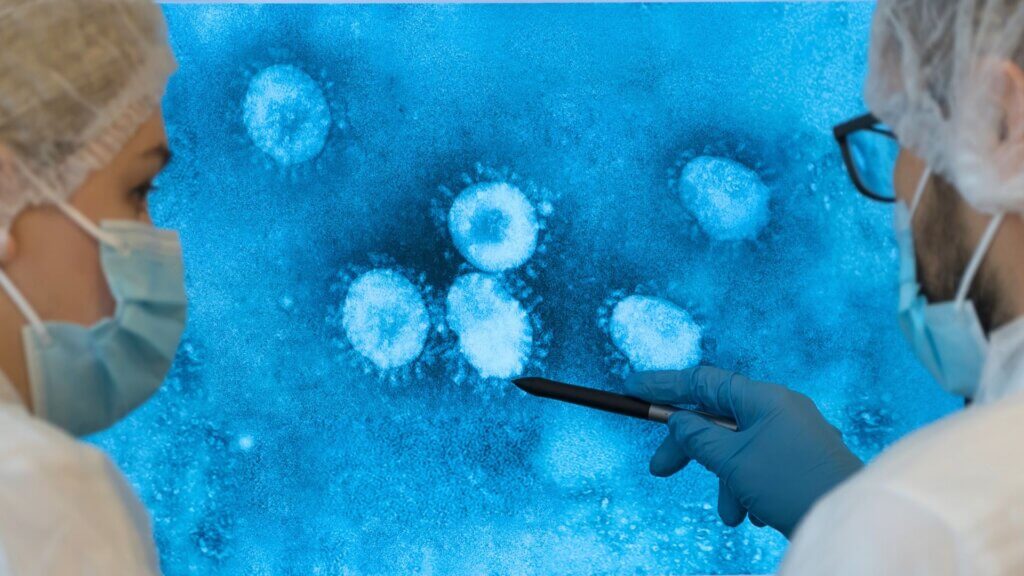 Scientists in protective masks and gloves examining a magnified image of virus particles on a laboratory screen.