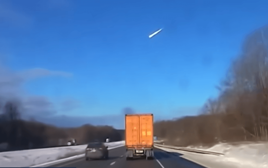 Dashcam view of a highway with a bright object streaking across the sky.