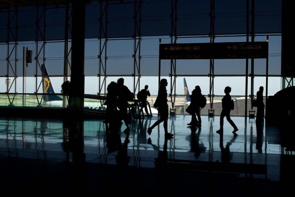 Silhouetted travelers walking through an airport terminal with planes visible outside.