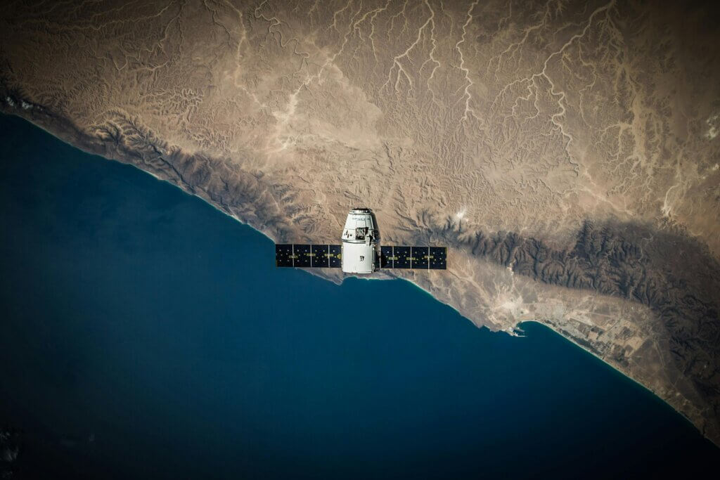 Space capsule with solar panels orbiting above Earth’s coastline.