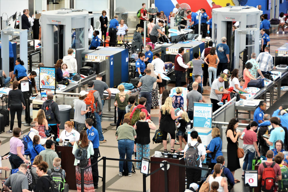 Travelers going through airport security screening lines with carry-on bags and TSA agents assisting passengers.
