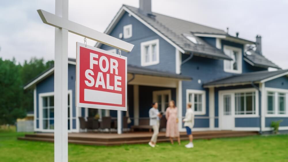 For sale sign in front of a suburban house as prospective buyers speak with a real estate agent.