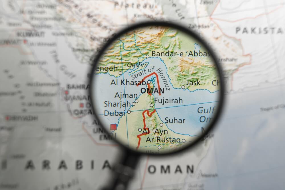 Magnifying glass highlighting the Strait of Hormuz between Iran, the UAE, and Oman on a regional map.