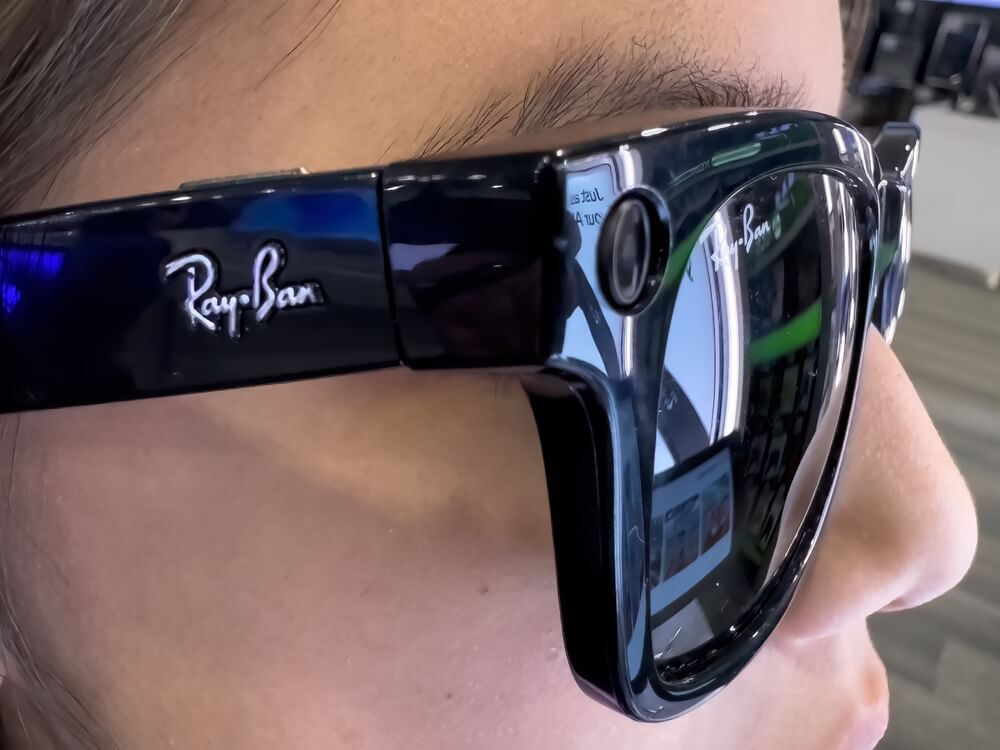 Person wearing black Ray-Ban Meta smart glasses with built-in camera lens near the hinge.
