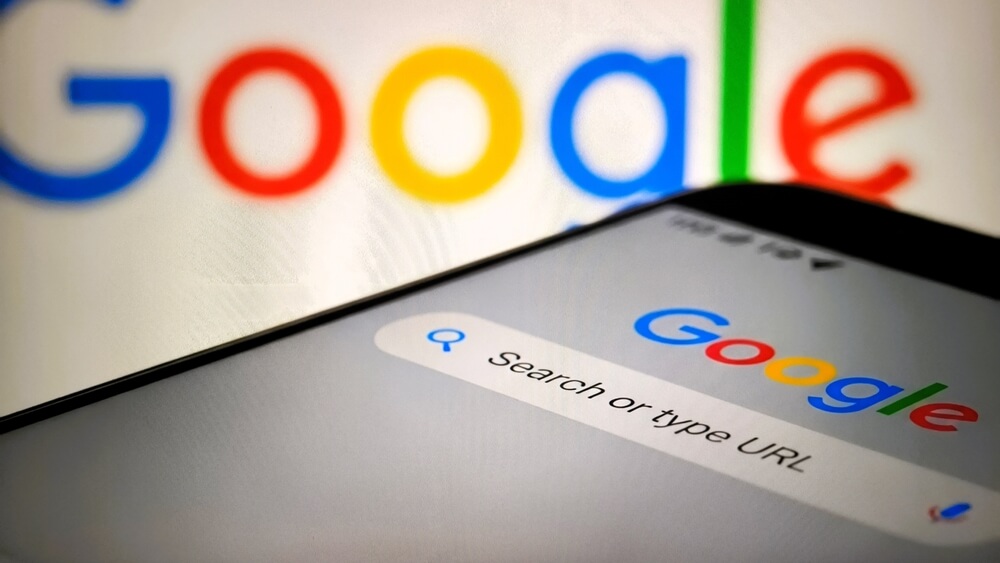 Smartphone showing the Google search bar and logo, with a blurred Google logo in the background.
