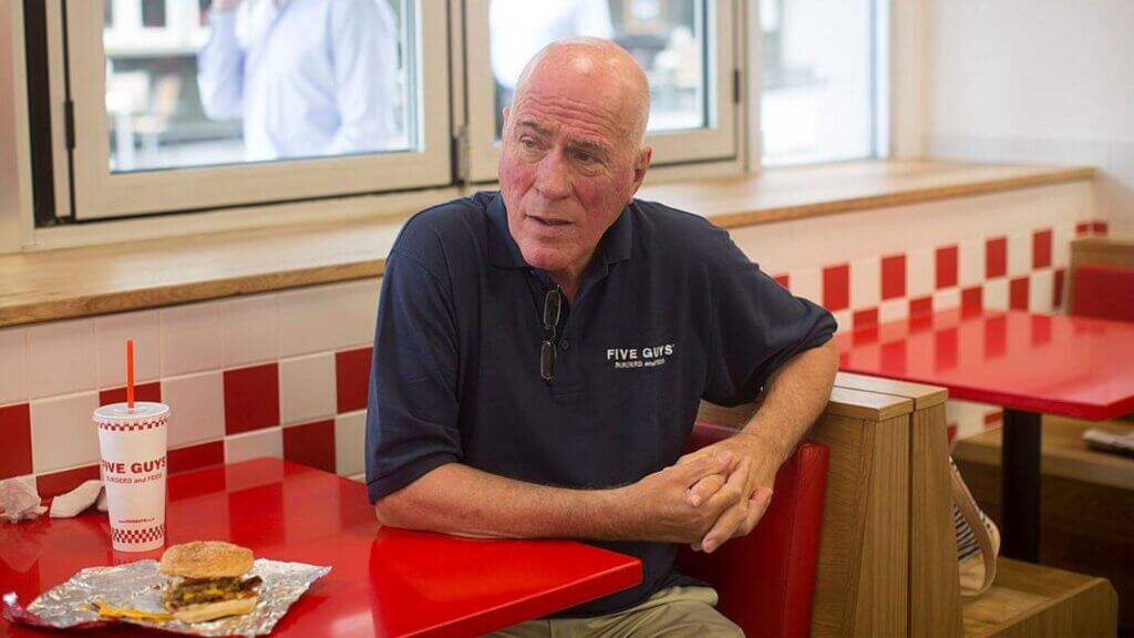 Five Guys CEO, Jerry Murrell, sitting at a booth with a burger and a drink on the table.