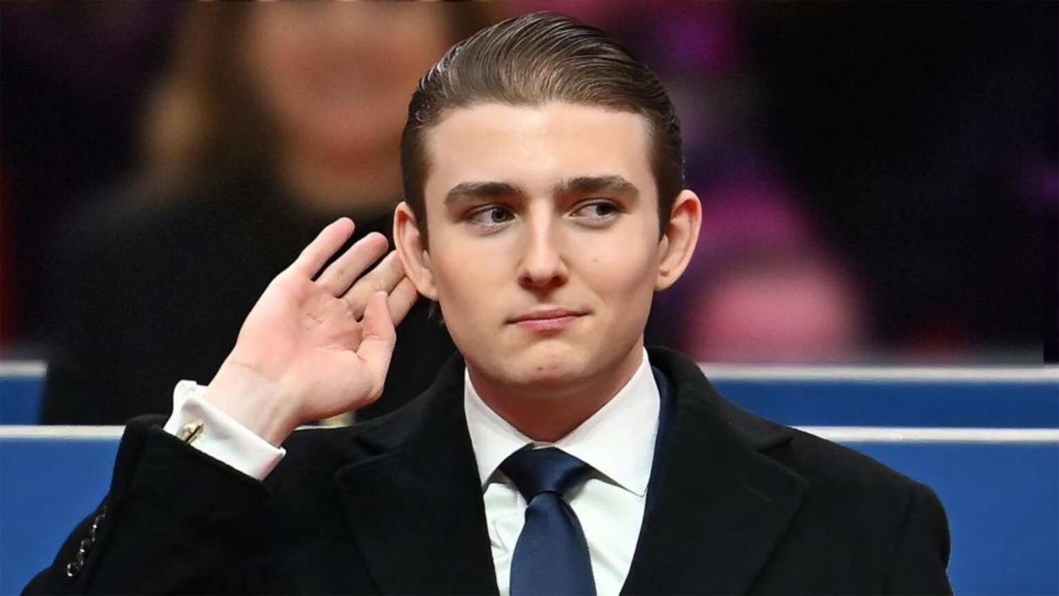 Barron Trump in suit raising hand to his ear at a formal event.