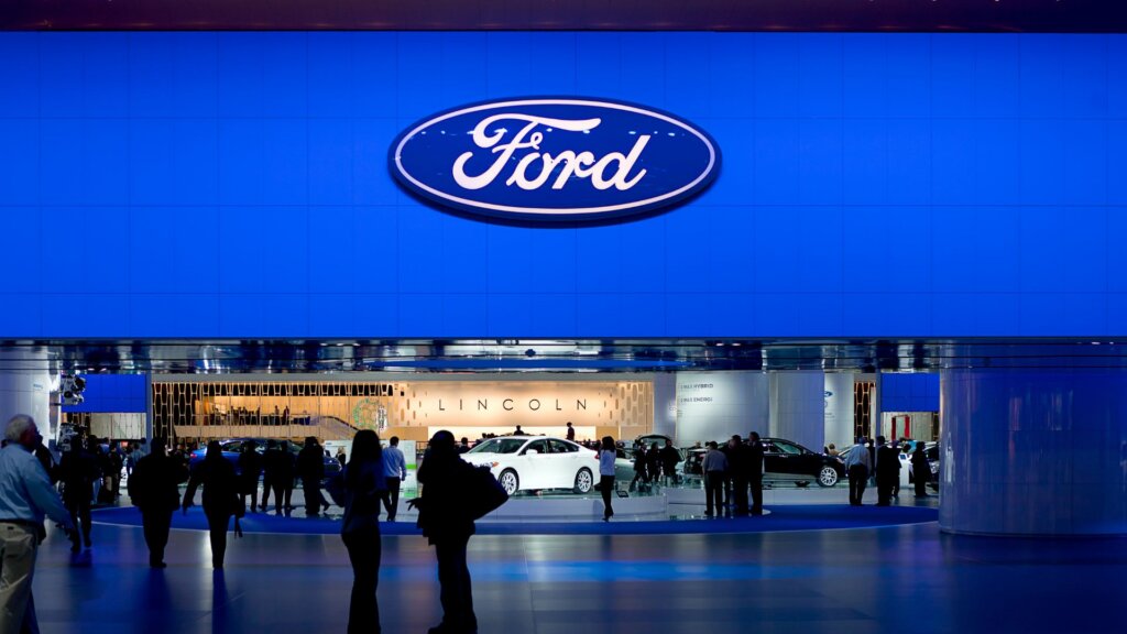 Ford logo above display of vehicles and crowd at an auto show booth.