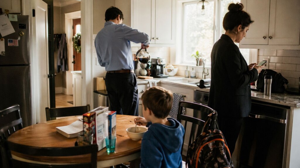 A family in a busy kitchen on a weekday morning, both adults preparing to leave for work while a child eats breakfast