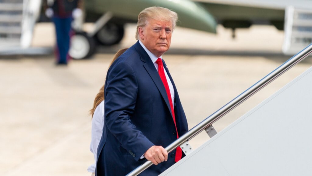Donald Trump in a navy suit with a red tie climbs the stairs to a plane while looking back toward the camera on an airport tarmac.