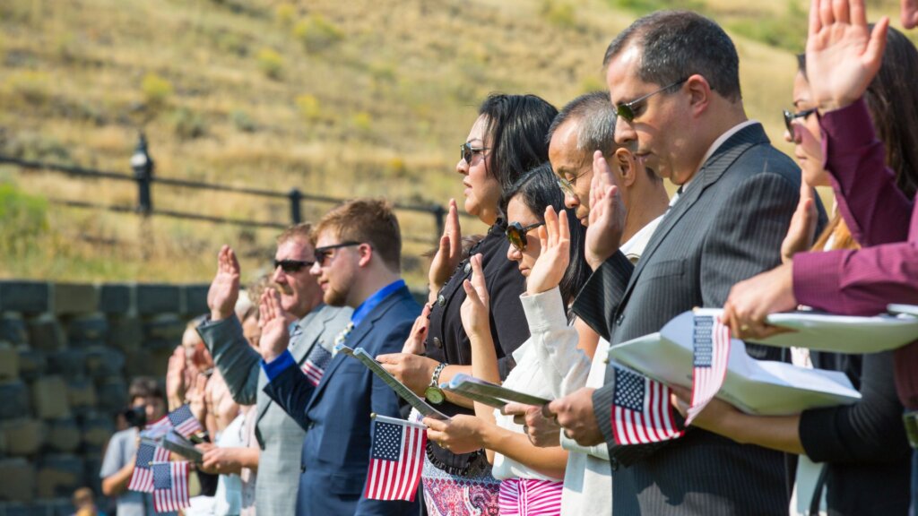 Immigrants raise hands taking oath of citizenship while holding U.S. flags at outdoor ceremony.