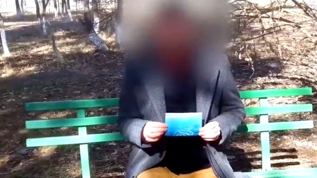 A medium shot of a man sitting on a green park bench in a wooded area; his face is heavily blurred, and he is wearing a dark grey blazer over a black shirt while holding a small, blue photograph in both hands.