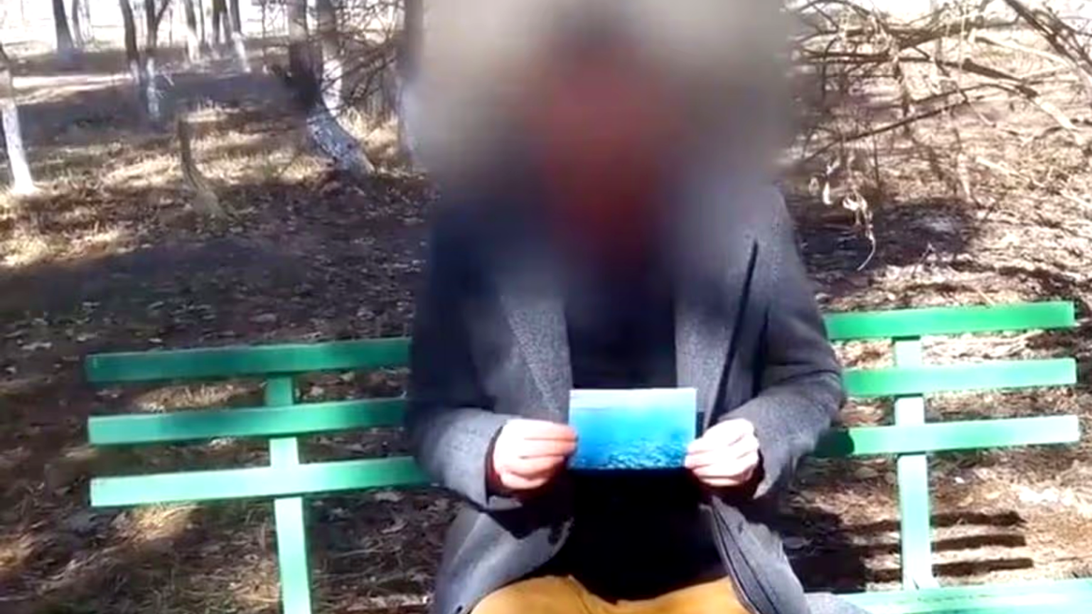 A medium shot of a man sitting on a green park bench in a wooded area; his face is heavily blurred, and he is wearing a dark grey blazer over a black shirt while holding a small, blue photograph in both hands.