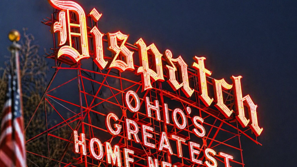 A low-angle shot of the historic neon "Dispatch" sign in Columbus, Ohio, glowing orange and white against a dark twilight sky.