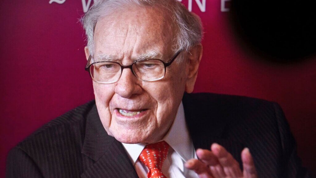 Warren Buffett speaking during an interview or event against a red backdrop.