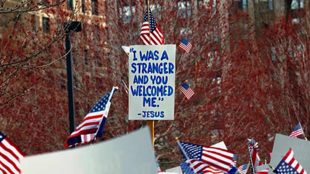 A protest scene showing a white sign with blue text held aloft amidst a sea of waving American flags in an urban setting.