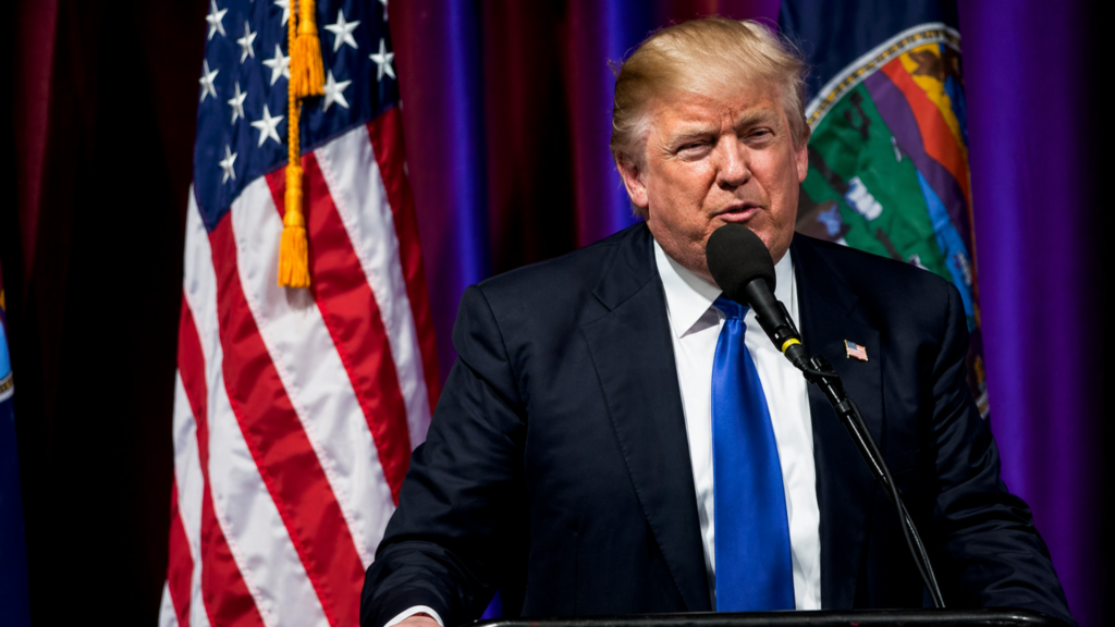 Donald Trump speaking at a podium with U.S. flag and state flag behind him during a public event.