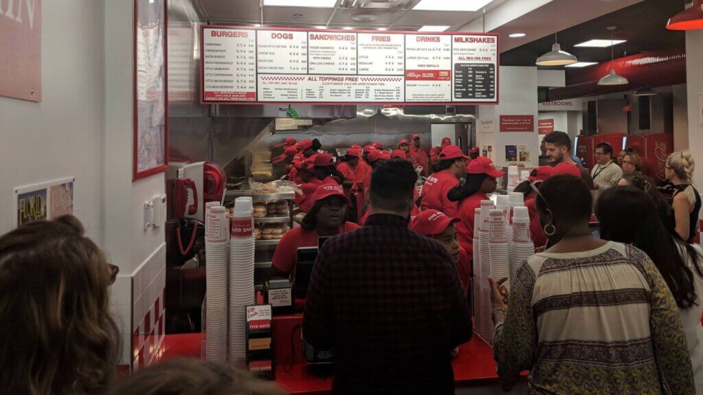 Customers ordering at crowded Five Guys counter with staff in red uniforms behind register.