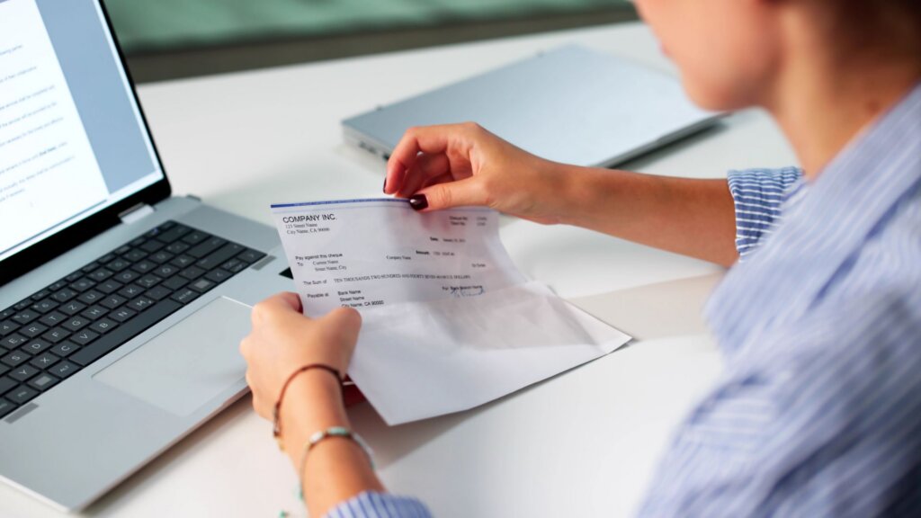 Woman opening a paycheck envelope beside a laptop at a desk.