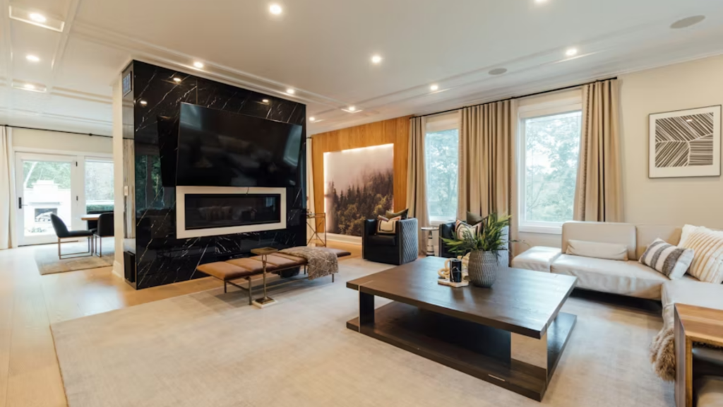 A wide interior shot of a minimalist, modern living room featuring a black marble wall with a fireplace and television, light wood floors, and a large windows overlooking a green forest.