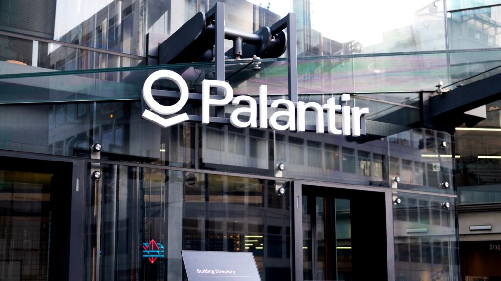 Palantir logo displayed on glass office building entrance in an urban setting.