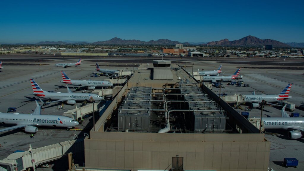 Multiple American Airlines planes are parked at gates at a large airport with desert mountains in the distance under a clear blue sky.