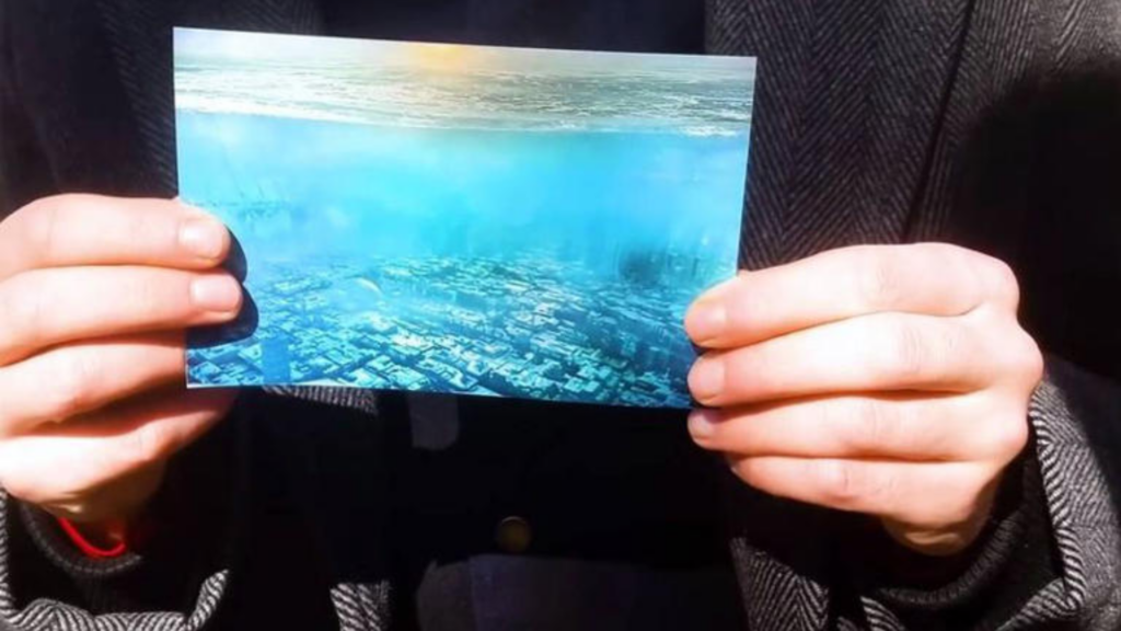 A close-up of a person's hands holding a rectangular photograph that appears to show a vast, futuristic city with skyscrapers submerged under clear, bright blue water, with a hazy sun visible at the top.