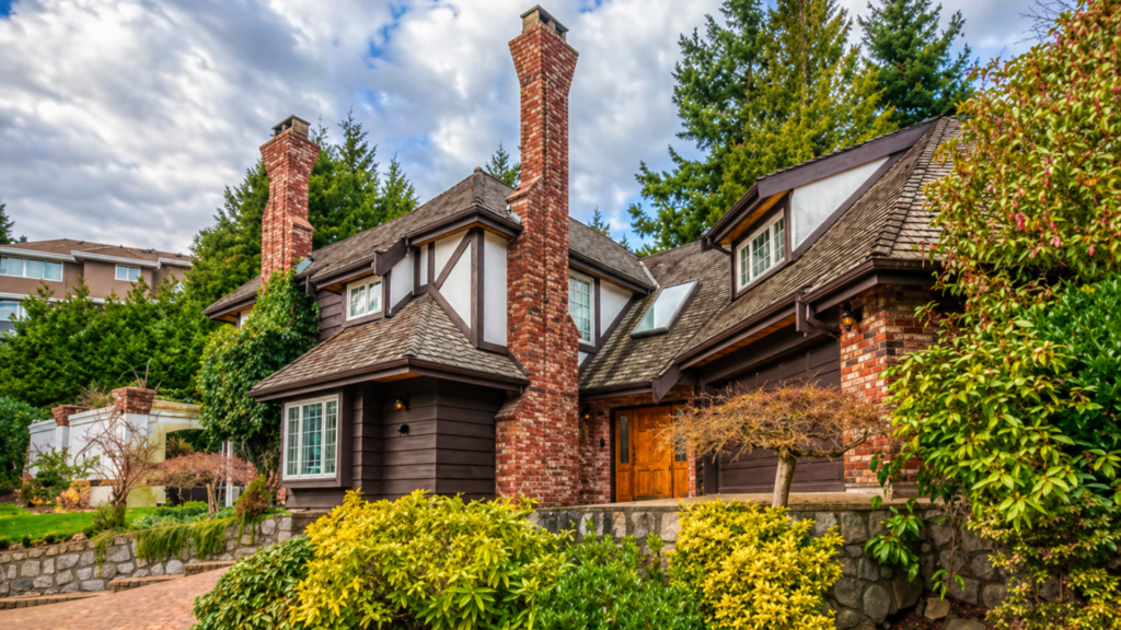 A professional photograph of a large, high-end suburban house with a dark brown wood and brick exterior, featuring multiple steep gables, two tall brick chimneys, and a multi-car garage surrounded by lush green trees and manicured yellow shrubs.