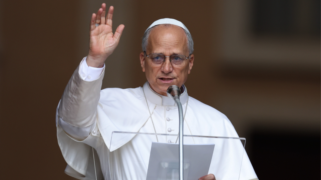 Pope Leo XIV speaking at a podium and raising his hand during a public address.
