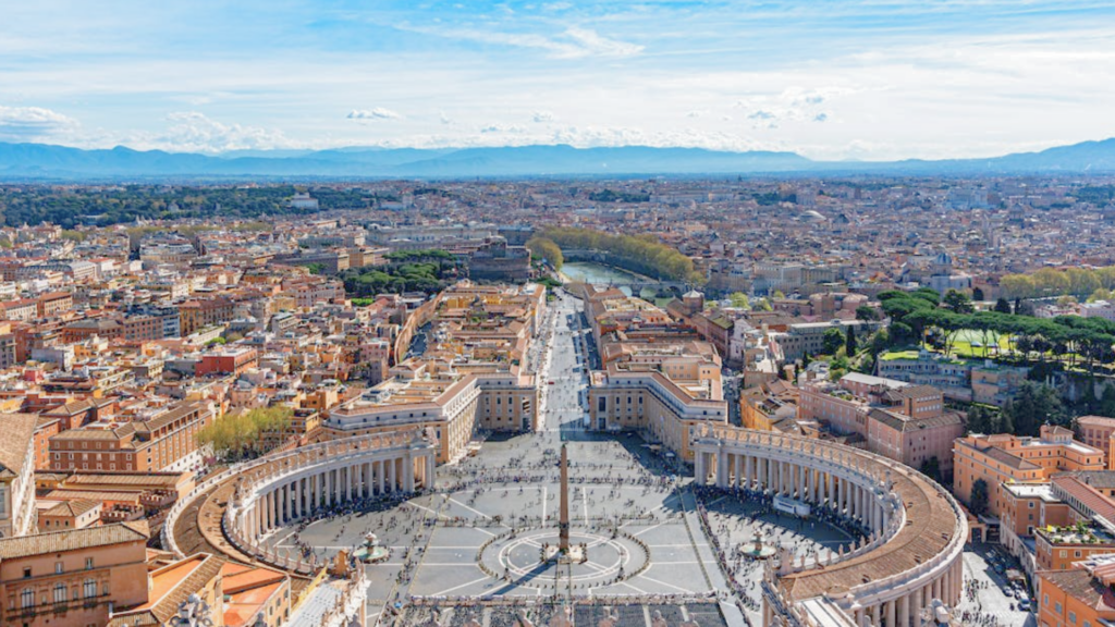 An aerial view looks down on the large, circular plaza of St. Peter's Square and the sprawling city of Rome.