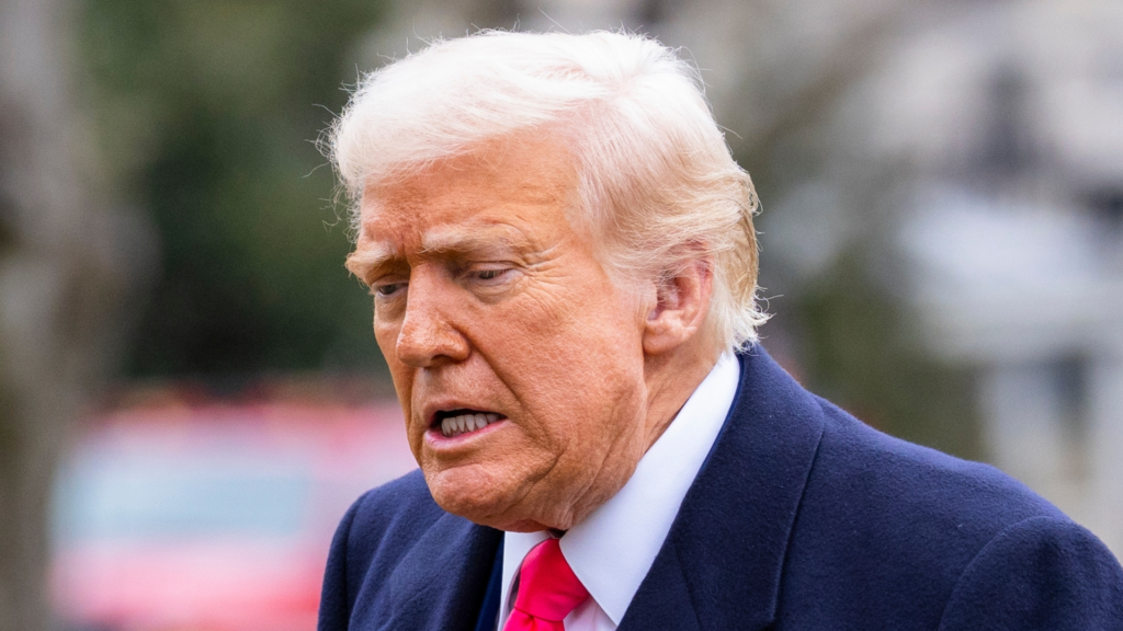 A close-up of Donald Trump looking downward with a serious expression, wearing a dark coat and a bright pink tie.