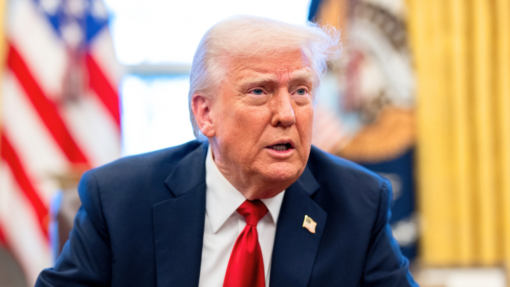 Donald Trump wears a dark suit and red tie while speaking in front of an American flag.