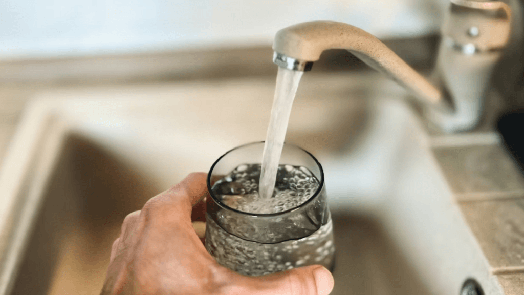 A hand holds a clear glass under a running faucet to fill it with water.
