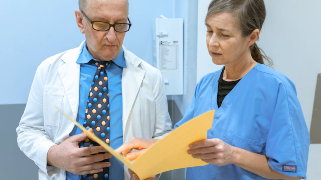 A doctor in a white coat and a nurse in blue scrubs look together at a yellow folder.