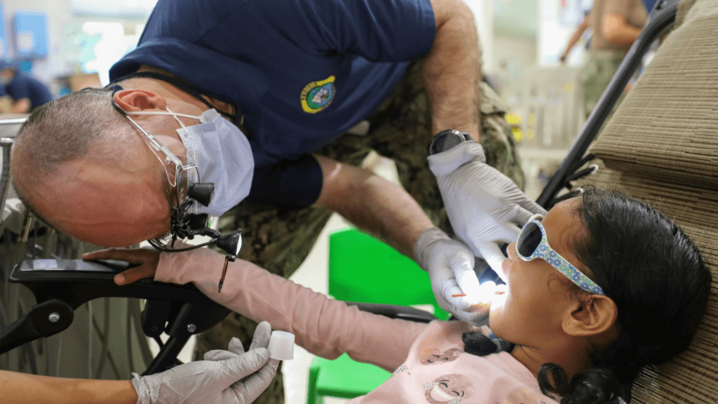 A dentist wearing a mask looks into the mouth of a young girl sitting in a chair.