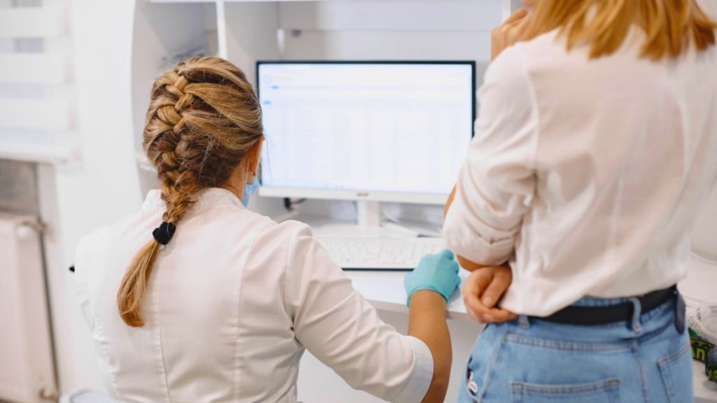 A healthcare worker wearing blue gloves works at a computer monitor while another woman stands nearby.